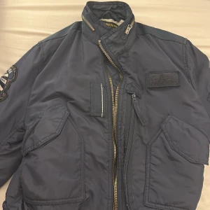 Mörkblå bomberjacka Alpha Industries - Säljer en mörkblå bomberjacka från Alpha Industries med coola patchar och detaljer. Jackan har flera fickor med dragkedja, ribbad krage och muddar samt snygga broderade märken på ärmen och bröstet. Perfekt för dig som gillar streetwear och militärinspirerad stil. Jackan är ej använd fick den som present av min vän då var den liten och därför har jag inte kunnat använda den så därför tänkte jag sälja den priset kan diskuteras vid snabb affär 