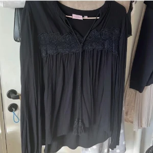 Svart blus med spets - Svart blus från KappAhl med lång ärm och dekorativ spets över bröstet. Blusen har en v-ringad hals med snörning och tofsar framtill. Materialet är mjukt och lätt, perfekt för dig som gillar en bohemisk vibe. Snyggt flowy fall och elastiska ärmslut.