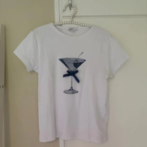 Vit baby tee med martiniglas från Subdued - Vit baby tee från Subdued med tryck av ett martiniglas och en blå rosett framtill. Klassisk rund halsringning och korta ärmar. Perfekt för dig som gillar coola och lekfulla prints.