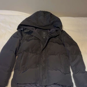 Svart pufferjacka från Canadian Goose - Svart pufferjacka från Canadian Goose med huva och emblem på ärmen. Jackan har quiltad design, dragkedja och knappar framtill. Perfekt för kalla dagar och har en snygg, modern look som funkar till streetwear. Passformen är normal och jackan är långärmad.