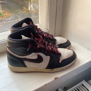 Nike Air Jordan 1 bloodline - Säljer ett par Nike Air Jordan 1 med svart och vitt skinn, röda detaljer och röda snören med text. Klassisk high-top siluett och perforerad tåbox. Snygg kontrast mellan färgerna och ikonisk Swoosh-logga på sidan. Perfekta för dig som vill sticka ut med dina sneakers.