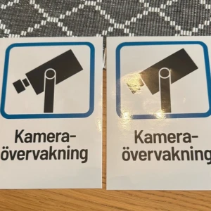 Kameraövervakning  - Två klistermärken med texten 'Kameraövervakning' och symbol för övervakningskamera. Perfekt för att informera om kameraövervakning i lokaler eller på tomt. Båda är helt nya!