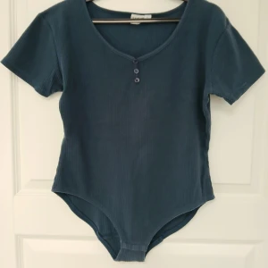 Body med knappar - En blå body med korta ärmar och tre tryckknappar i grenen. Tillverkad i mjuk bomull (95%) och elastisk elastan (5%). Har tre dekorativa knappar (en extraknapp medföljer) framtill. Perfekt för att styla med t.ex. kjol för en chill look. Plagget har naturligtvis tvättats. Se bilder för skick och passform. Armhåla till armhåla är cirka 47 cm. Axelsöm till ärmslut är cirka 15 cm. Tvättas i 40 grader.