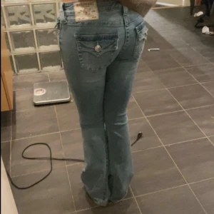 Ljusblå bootcut jeans från True Religion - Säljer ett par ljusblå jeans från True Religion med bootcut passform. Jeansen har låg midja, klassiska silverfärgade knappar och detaljerade bakfickor med lock och knapp. Tillverkade i jeansmaterial med snygg tvätt och ikonisk etikett bak.