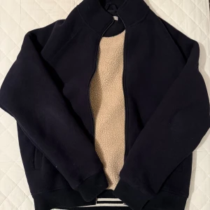 Svart teddyjacka  - Svart teddyjacka från MNG Man i storlek M. Jackan har mjukt beige teddyfoder, ribbade muddar och krage, samt dragkedja framtill. Snygga svarta detaljer och randig kant längst ner. Perfekt för kalla dagar när du vill vara både chill och varm.