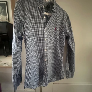 Rutig blå skjorta Polo Ralph Lauren - Säljer en klassisk blå och vit rutig skjorta från Polo Ralph Lauren i slim fit. Skjortan har långa ärmar, knappar framtill och den ikoniska broderade loggan på bröstet. Perfekt för dig som gillar stilrena och tidlösa plagg.