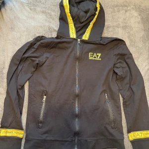 Svart EA7 hoodie med gula detaljer - Svart hoodie från EA7 Emporio Armani med dragkedja, huva och snygga gula logodetaljer på huva och ärmslut. Tröjan har två fickor med dragkedja och EA7-tryck på bröstet. Perfekt för dig som gillar streetwear och vill sticka ut.