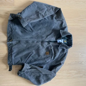 Grå workwear jacka från Carhartt - Säljer en grå workwear jacka från Carhartt med klassisk krage, dragkedja framtill och två snedställda fickor. Jackan har robust bomullsmaterial och detaljer med tryckknappar vid ärmslut och midja. Perfekt för dig som gillar streetwear och funktionell stil.