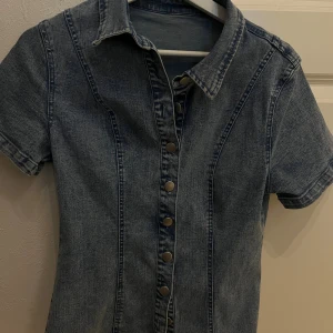 Jeanströja med knappar - Säljer en jeanströja i klassisk blå denim med korta ärmar och krage. Tröjan har snygga silverfärgade tryckknappar framtill och markerade sömmar som ger en cool och figurnära look. Perfekt för dig som gillar enkel och stilren denim.