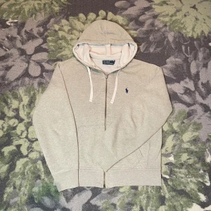 Ljusgrå hoodie från Polo Ralph Lauren - Ljusgrå hoodie från Polo Ralph Lauren med dragkedja och huva med vita snören. Klassisk blå logga broderad på bröstet. Tillverkad i mjuk bomull med ribbade muddar och fickor framtill. Perfekt för chill dagar och enkel att matcha med jeans storlek S.