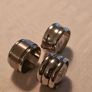 Breda silverfärgade ringar storlek 19 - Tre breda ringar i silverfärg med stilren och modern design. Två av ringarna har en slät yta med en diskret upphöjd kant, medan den tredje har tre markerade ränder runt om. Alla är I Rostfritt stål och ca 19 mm