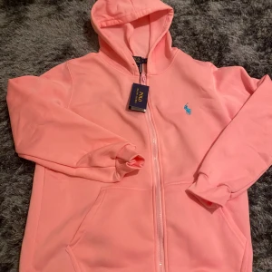 Rosa hoodie från Polo Ralph Lauren - Snygg rosa hoodie från Polo Ralph Lauren med dragkedja och huva. Tröjan har ett blått broderat logomärke på bröstet, ribbade muddar och två fickor framtill. Tillverkad i mjuk bomull med stretch för extra komfort. Perfekt för chill dagar.