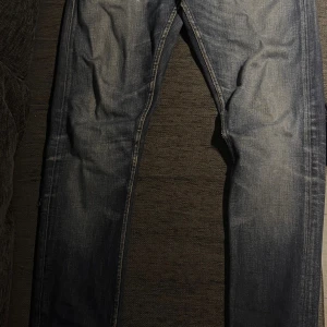 Blå jeans med slitningar - Säljer ett par blå jeans med snyggt slitna detaljer och ljusare partier på låren och knäna. Klassisk femficksmodell med orange sömmar och dragkedja. Jeansen har en rak passform och är tillverkade i robust denim. Litet hål i byxorna där back men kan lätt sys ihop. märke Tiger of Sweden