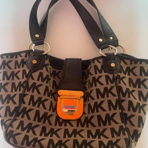 MK axelväska med monogram och detaljer - Snygg axelväska från Michael Kors med MK-monogram över hela väskan i svart mot beige bakgrund. Väskan har svarta handtag i skinn, guldfärgade metalldetaljer och ett coolt lås framtill. Perfekt storlek för allt du behöver och riktigt trendig look.