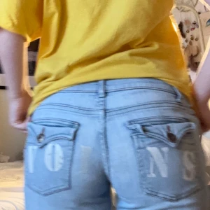 Ljusblå bootcut jeans med rå kant - Säljer ett par ljusblå jeans med bootcut passform och rå, fransig kant nertill. Jeansen har låg midja, klassiska fickor med lock och tryckknappar bak samt tryckta bokstäver på bakfickorna. Tillverkade i jeansmaterial och perfekta för en avslappnad stil. De passar mig som är 163 cm och brukar bära M. 