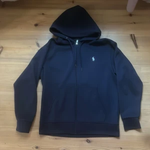 Marinblå hoodie från Polo Ralph Lauren - Säljer en marinblå hoodie från Polo Ralph Lauren med dragkedja och klassisk logga broderad på bröstet. Tröjan har huva med snörning, två fickor fram och är tillverkad i mjuk bomullsblandning. Perfekt för en chill och stilren look. 