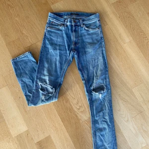 Blå slitna jeans från Nudie Jeans - Säljer ett par blå jeans från Nudie Jeans med slitningar och hål på båda knäna. Klassisk femficksmodell med orangea sömmar och broderad detalj på bakfickorna. Jeansen har en rak passform och är tillverkade i bomull. Längd 30 waist 27