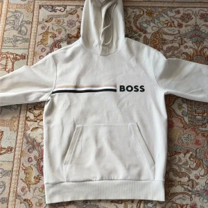 Vit hoodie från BOSS med tryck - Snygg vit hoodie från BOSS med svart logga och stilrena ränder i svart, vitt och beige över bröstet. Klassisk känguruficka framtill, huva med dragsko och mjukt bomullsmaterial. Perfekt för en chill och clean look.