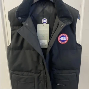 Svart dunväst från Canada Goose - Svart dunväst från Canada Goose med klassisk logga på bröstet, hög krage och stora fickor framtill. Västen har både dragkedja och tryckknappar samt quiltad design. Perfekt för lager-på-lager och riktigt snygg till streetwear.