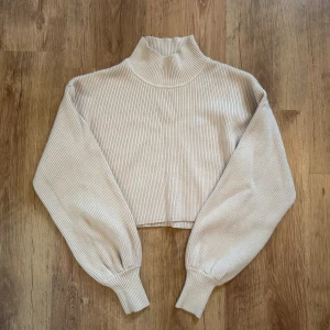 Fin Beige croppad polotröja Gina Tricot - Snygg croppad polotröja från Gina Tricot i beige med ribbad struktur. Tröjan har lång ärm med lätt puff och hög krage. Materialet är en mix av viskos, polyester och polyamid som ger en mjuk och skön känsla. Perfekt för dig som gillar trendiga och bekväma plagg.