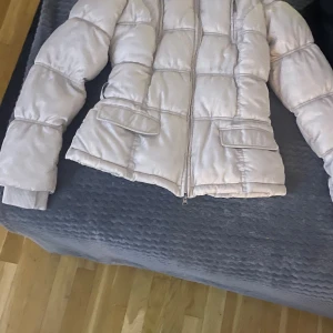 Vit pufferjacka från Gaudi Jeans - Säljer en vit pufferjacka från Gaudi Jeans med hög krage och två stora fickor framtill. Jackan har quiltad design, dragkedja och ribbade muddar vid ärmslut. Perfekt för kalla dagar och har en clean, modern look.