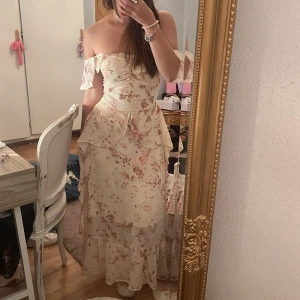 Blommig offshoulder långklänning från LAM - Romantisk offshoulder långklänning från LAM med volangdetaljer och blommigt mönster i ljusa toner av beige och rosa. Klänningen har axelbandslös design, omlottliknande överdel och mjukt, luftigt material som ger en flowy känsla. Perfekt för sommarens alla tillfällen.
