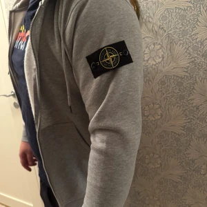 Grå hoodie från Stone Island - Säljer en grå hoodie från Stone Island med dragkedja, huva och snörning. Klassisk logga på vänster ärm och två fickor framtill. Jättefint skick, inga defekter. 