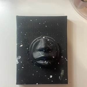 Modern Tavla - Ingen elektronikprodukt identifierad på bilderna. Objektet är en konstnärlig tavla med tredimensionell ansiktsform, målad i svart med vita stänk.  inget märke eller modell.