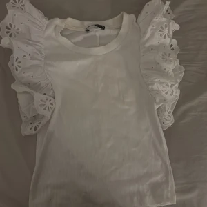 Vit tröja med volangärmar  - Säljer en vit tröja med ribbad kropp och stora volangärmar i broderad bomull med blommönster. Ärmarna är korta och har snygga hålbroderier som ger en cool och romantisk vibe. Perfekt för dig som vill sticka ut med detaljerade ärmar.