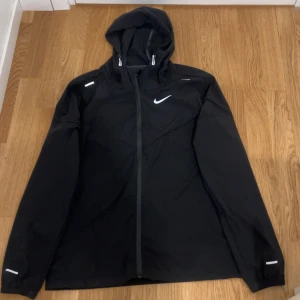 Svart Nike vindjacka med huva - Svart vindjacka från Nike med huva och reflekterande detaljer på axlar och ärmar. Jackan har dragkedja framtill och en stilren design med Nike-logga på bröstet. Perfekt för träning utomhus när det blåser. Långärmad och lätt material.