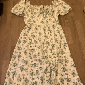 Vit blommig långklänning med slits - Är i L men passar mer S/M Superfin långklänning i vit med grönt blommönster. Klänningen har korta puffärmar, slits framtill och snörning vid bysten. Perfekt för sommarens alla tillfällen och ger en romantisk vibe. Från SHEIN tyvärr 
