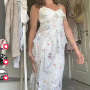 Vit långklänning med blommigt mönster -  vit långklänning med tunna axelband och blommigt mönster i pastellfärger. Klänningen har volangdetaljer och omlottkänsla framtill, vilket ger en luftig och somrig look. Perfekt för varma dagar och speciella tillfällen.