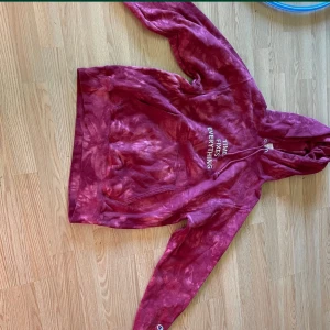 Röd tie-dye hoodie från Champion - Säljer en röd tie-dye hoodie från Champion med texten 'TIME FIXES EVERYTHING' på bröstet. Tröjan har huva med snörning, magficka och långa ärmar. Perfekt för dig som gillar streetwear och vill sticka ut lite extra.