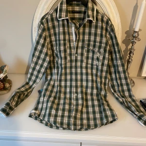 Grön rutig skjorta från Sisley - Snygg grön och beige rutig skjorta från Sisley med vit kant på kragen. Skjortan har två bröstfickor med knapp, klassisk krage och knappar framtill. Perfekt för dig som gillar en chill och stilren look. Materialet är mjuk bomull som känns skönt mot huden.