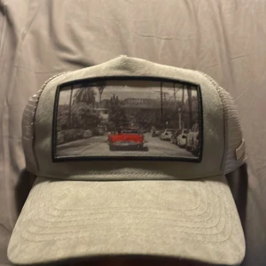 Grå keps från Lxery med mesh och tryck - Snygg grå keps från Lxery med mesh på sidorna och bak för extra ventilation. Framtill finns ett coolt tryck med en röd bil på en stadsgata, vilket ger kepsen en unik vibe. Perfekt för dig som vill sticka ut med en urban stil. Skriv vid minsta fundering. Pris kan diskuteras 