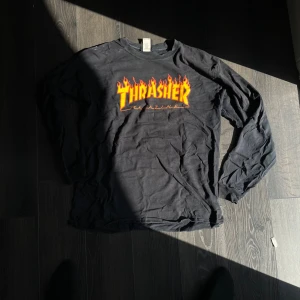 Svart Thrasher långärmad tröja - Svart långärmad Thrasher-tröja med ikoniskt logotryck i gult och rött med eldflammor på bröstet. Skön passform och perfekt för dig som gillar streetwear och skatekultur. Tillverkad i mjuk bomull.