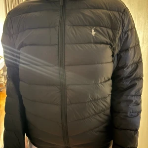 Svart pufferjacka från Polo Ralph Lauren - Svart pufferjacka från Polo Ralph Lauren med huva och dragkedja framtill. Jackan har quiltad design, två sidofickor med dragkedja och den klassiska Polo-loggan broderad på bröstet. Perfekt för kalla dagar och riktigt snygg streetstil.