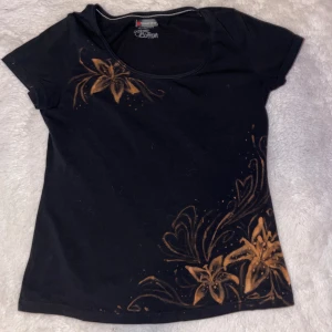 Svart blommig t-shirt  - Svart t-shirt från Street One i ekologisk bomull med orangea blommor och blad som mönster framtill. Har själv blekt mönstret. Modellen har rund halsringning och korta ärmar, sitter tight och har ett mjukt material som känns skönt mot huden. Perfekt för dig som gillar unika prints och chill stil.