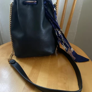 Svart skinnväska & Other Stories  - Stilren svart bucket bag i skinn med guldfärgade metalldetaljer och kedjerem. Väskan har snörning med ett dekorativt blått mönstrat band och en stadig botten med metallfötter. Perfekt för dig som vill ha en trendig och rymlig väska med coola detaljer.