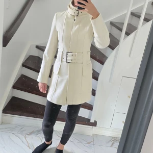 Elegant krämfärgad kappa med bälte – figursydd och stilren - Superfin kappa i krämvit färg med markerad midja och stort bälte som framhäver siluetten. Klassisk, tidlös modell som passar både till vardag och mer uppklädda tillfällen. Sparsamt använd.  Perfekt höst- och vinterkappa– stilren, varm och lätt att matcha! 