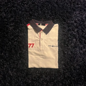 Vit Sail Racing pikétröja med tryck - Vit pikétröja från Sail Racing med mörkblå krage och röda detaljer. Snyggt tryck med siffran 77 på bröstet och Sail Racing-logga. Klassisk knappslå framtill och kontrastfärgade inslag vid kragen. Perfekt för dig som gillar sportig stil.