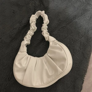 Beige axelväska med rynkad rem - Trendig beige axelväska i syntetmaterial med snygg rynkad axelrem och rundad form. Väskan har ett stilrent och minimalistiskt uttryck, perfekt för att lyfta din outfit. Rymligt huvudfack som stängs upptill.