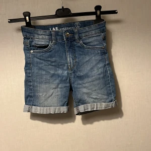 Blå jeansshorts från LAB Industries - Snygga blå jeansshorts från LAB Industries med klassisk femficksdesign och uppvikta benslut. Shortsen har normal passform och är tillverkade i slitstark bomull, perfekta för sommardagar. Stängs med knapp och dragkedja framtill.