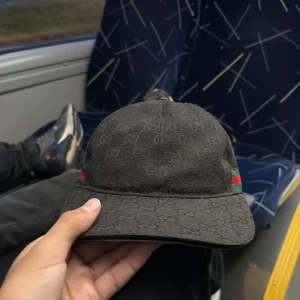 Svart Gucci keps med GG-mönster - Säljer en svart keps från Gucci med klassiskt GG-monogram över hela kepsen. Sidorna har gröna och röda ränder som signaturdetalj. Justerbar rem baktill i skinn. Snygg och stilren keps som lyfter vilken outfit som helst.