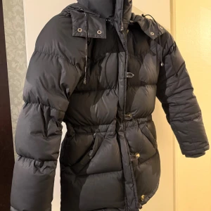 Svart Boomerang dunjacka med huva - Säljer en svart Boomerang dunjacka med huva och snörning. Jackan har långa ärmar, dragkedja och knappar framtill samt praktiska fickor. Puffad design som håller dig varm under kalla dagar. Perfekt för dig som vill ha en stilren och funktionell vinterjacka.