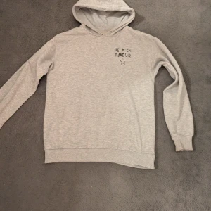 Grå hoodie med tryck Je m'en amour - Snygg ljusgrå hoodie med huva och lång ärm. På bröstet och ryggen finns ett svart tryck med texten 'Je m'en amour' och en stjärna. Tröjan har ribbade muddar och är gjord i mjuk bomull.