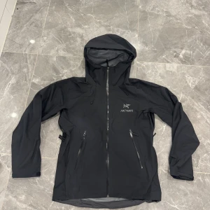 Svart Arc'teryx Gore-Tex vindjacka - Svart vindjacka från Arc'teryx med Gore-Tex-material, perfekt för blåsiga och regniga dagar. Jackan har huva, hel dragkedja framtill och två snedställda fickor med dragkedja. Snygg logga på bröstet och diskret Gore-Tex-brodyr på ärmen.