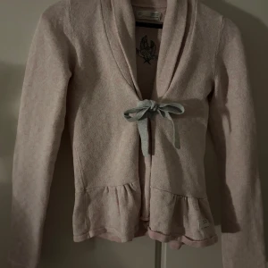 Ljusrosa kofta Odd Molly med rosett - Superfin ljusrosa kofta från Odd Molly med grå rosett framtill och volangdetaljer vid nederkanten. Långärmad och mjuk i materialet, med broderad Odd Molly-logga på ryggen. Perfekt för dig som gillar söta och feminina detaljer.