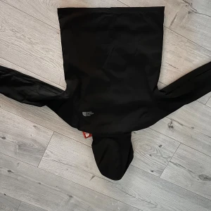 Svart vindjacka från The North Face - Svart vindjacka från The North Face med huva och dragkedja framtill. Jackan har två sidofickor med dragkedja och diskret logga på bröstet. Helt ny men passade inte mig jackan är en replicka därav det billiga priset.