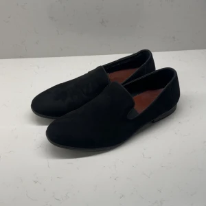 Svarta mockaloafers från Iguassu - Snygga svarta loafers från Iguassu Trade med mjuk mocka på ovansidan och diskret rund tå. Skorna har en klassisk slip-on design och innersulan är i brunt skinn med röd söm. Perfekta för dig som gillar stilrena och bekväma skor.